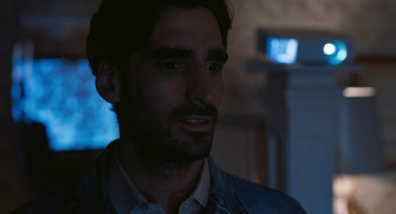 Matthias & Maxime (2019) movie still: closeup — A dimly lit indoor space is illuminated by a projected blue light source. A man in a denim jacket…