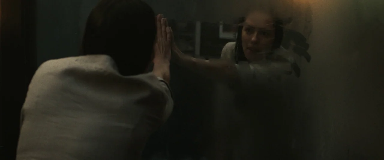 The Night House (2020) movie still: medium — A dimly lit room with a large wall-mounted mirror showing a reflection. A woman in a light shirt…