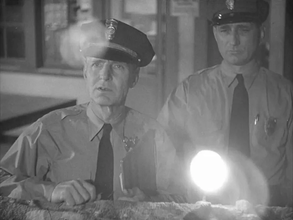 The Grapes of Wrath (1940) movie still: medium — Two police officers stand at a desk inside a dimly lit station interior. The frame is dominated by…