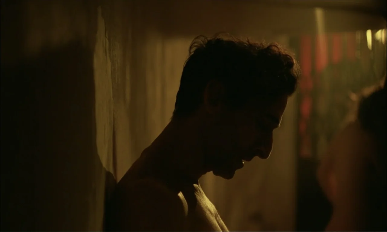 The Brutalist (2024) movie still: closeup — A dimly lit room features a man leaning against a textured plaster wall. The subject stands in profile…