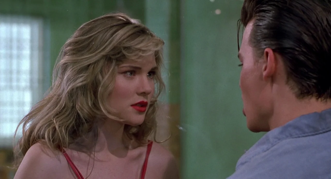Cry Baby (1990) movie still: medium — A dimly lit room with sage green walls and a small barred window nearby. A blonde woman in a red top faces a…
