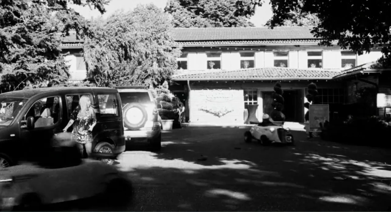 Do Not Expect Too Much from the End of the World (2023) movie still: wide — A paved lot in front of a low-rise building with mature trees overhead.…