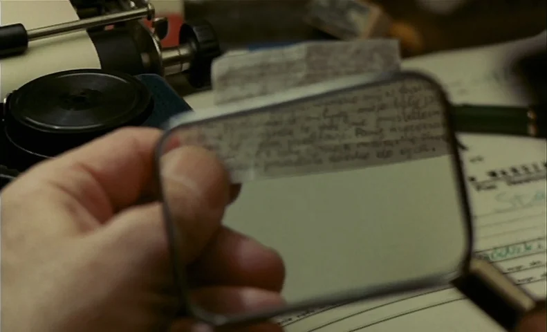No End (1985) movie still: closeup — A wooden desktop surface holds a vintage typewriter and a handwritten note. A person's hand uses a magnifying…