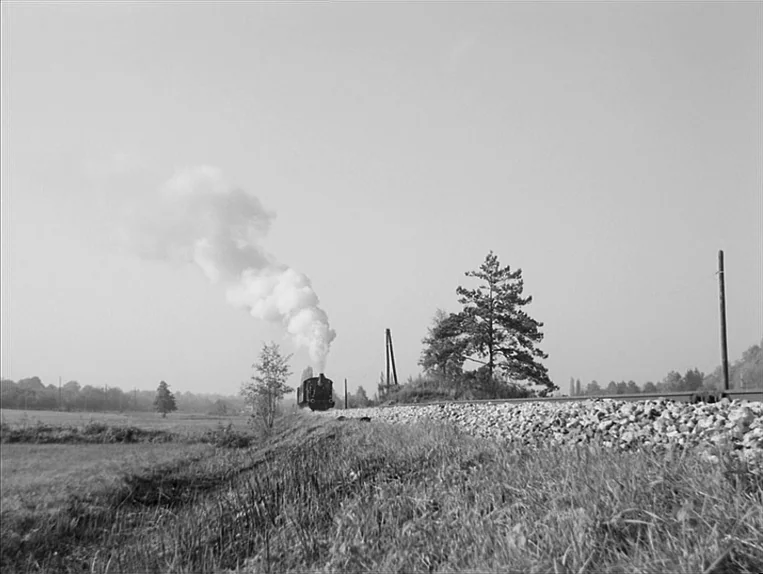 Effi Briest (1974) movie still: wide — A rural railway track stretches across a grassy field under a bright, overcast sky. A steam engine moves…