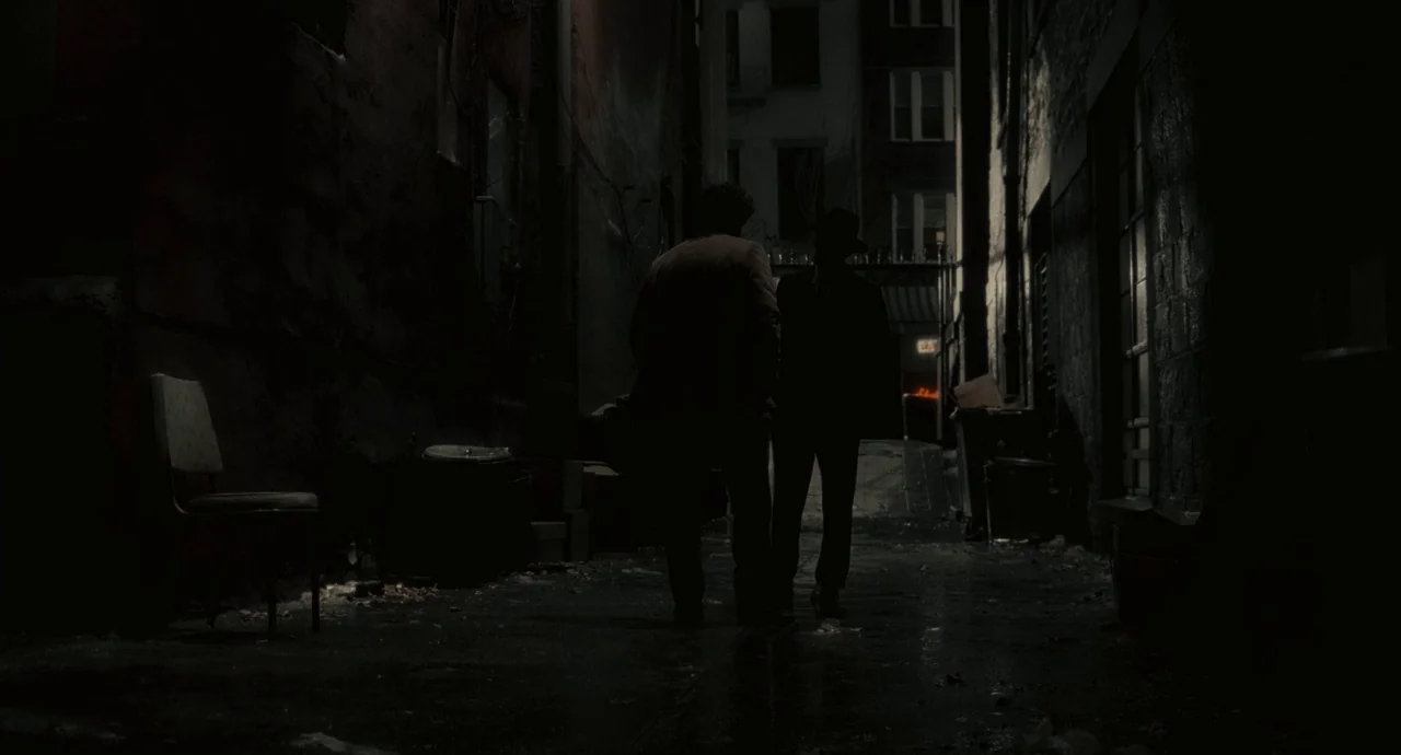Inside Llewyn Davis (2013) movie still: fullbody — A dark narrow urban passage with brick walls and a solitary chair left outside. Two men stand in…