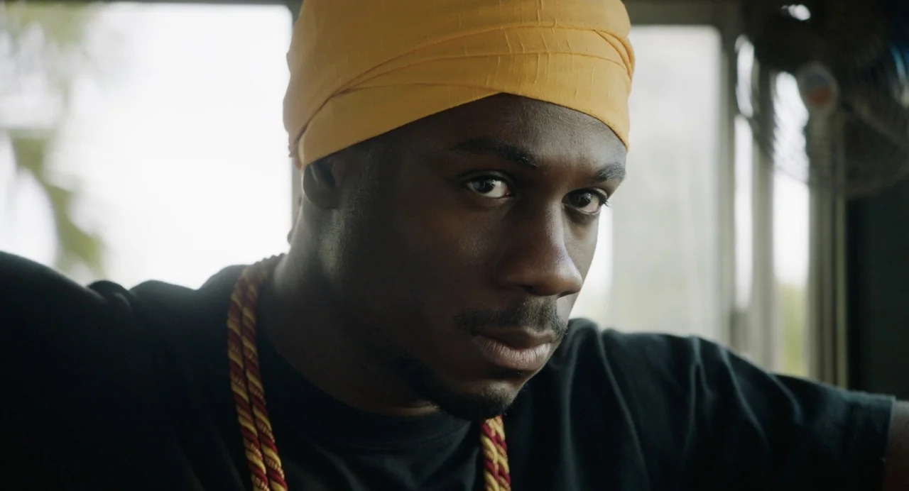 The Day Shall Come (2019) movie still: closeup — The interior of a vehicle shows a man positioned near a window. He looks directly forward while…