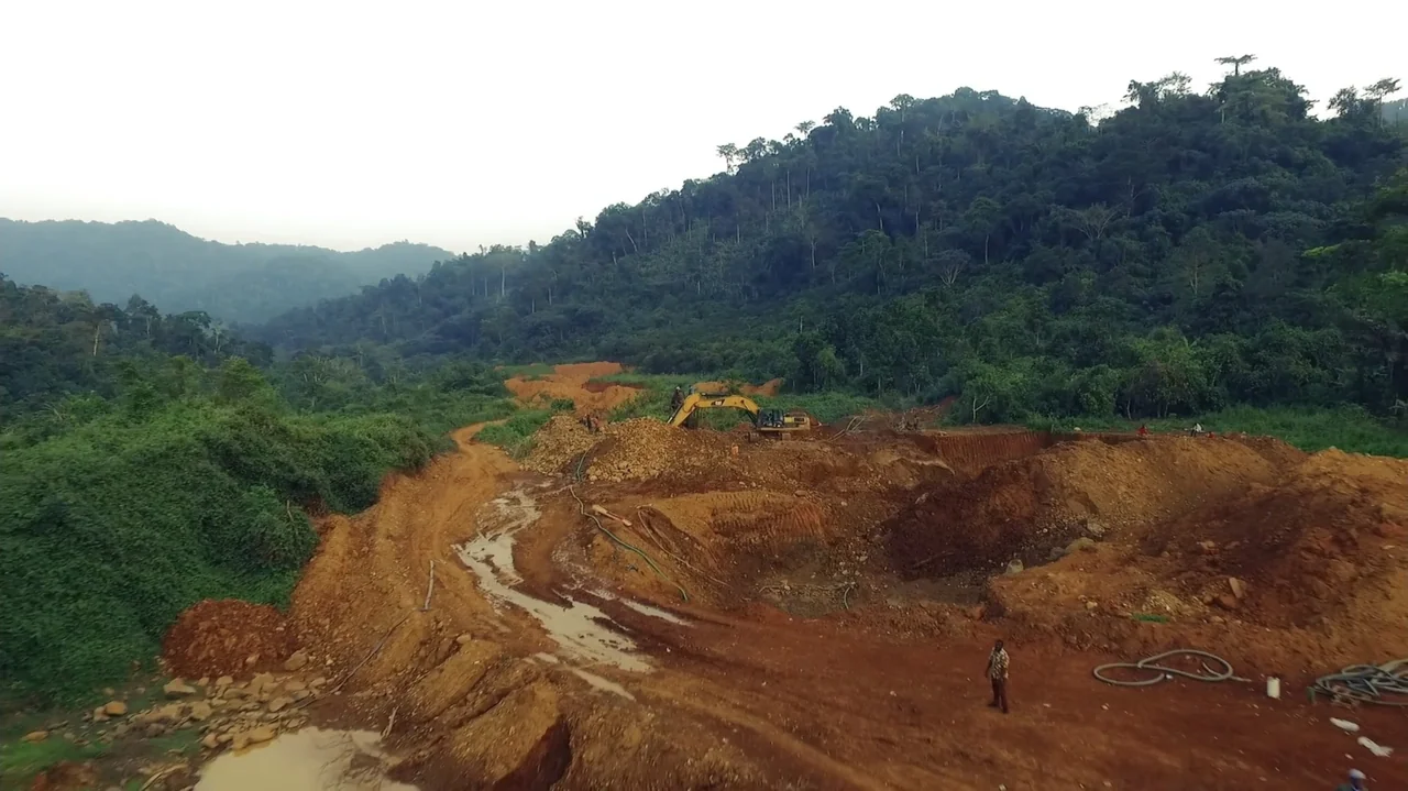 The Money Stone (2018) movie still: wide — A cleared patch of tropical hillside is undergoing excavation in a dense forest area. A yellow excavator…