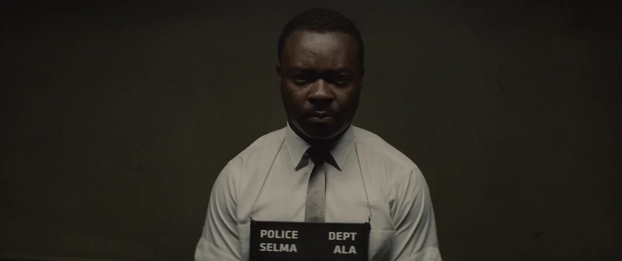 Selma (2014) movie still: medium — A man stands against a dark, plain wall inside a police facility. He wears a dress shirt and tie while holding a…