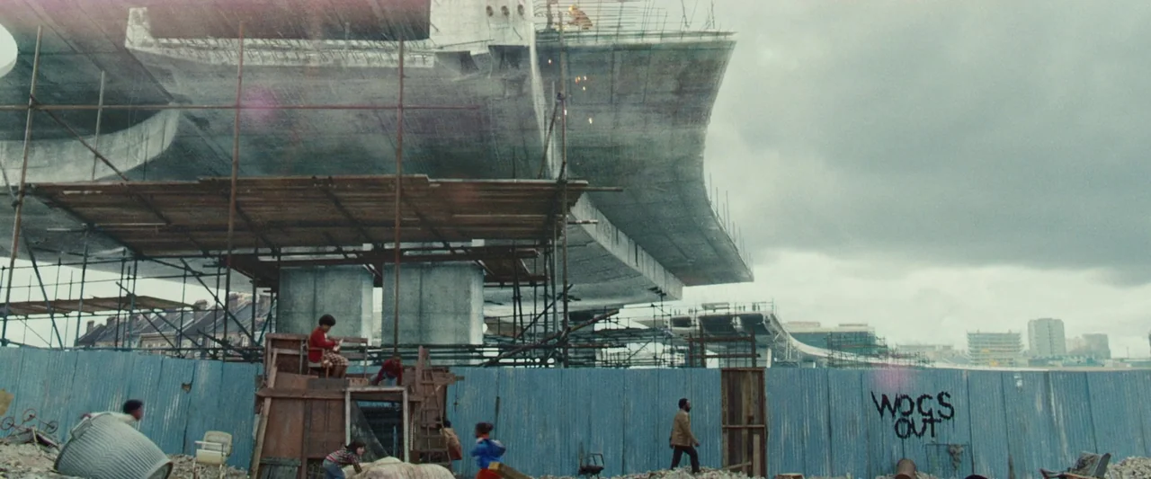 Mangrove (2020) movie still: wide — An urban construction site features a large elevated concrete highway section and scaffolding. Children play on…