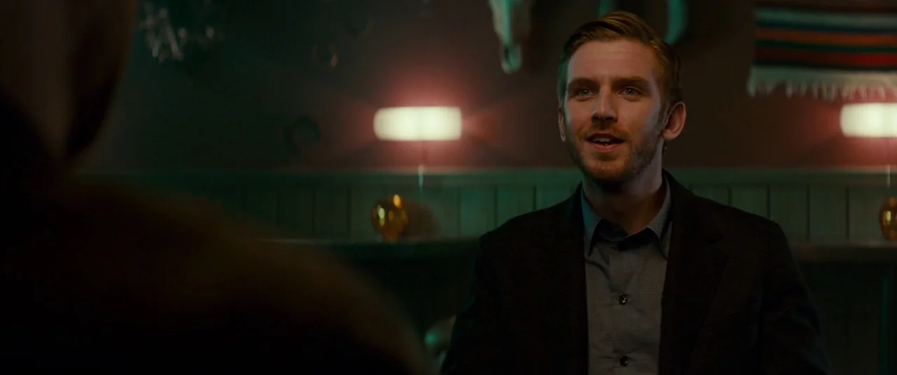 Colossal (2016) movie still: medium — A dark booth at night with two table lamps providing dim, warm light. A man in a dark suit sits in the…