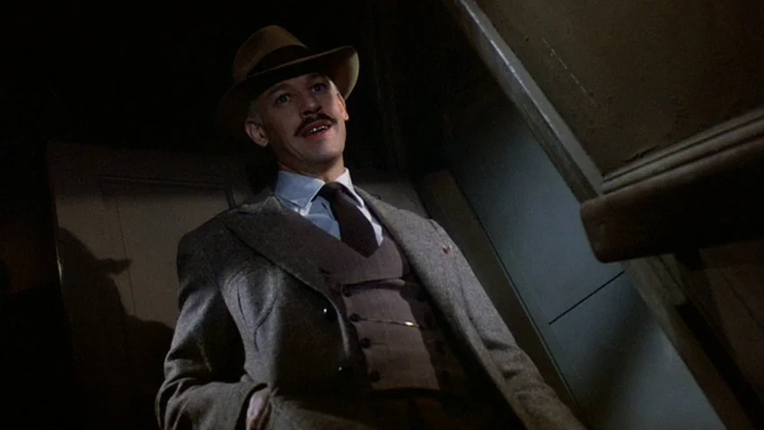 Hammett (1982) movie still: medium — A man stands on a staircase in a dark, narrow domestic interior setting. He looks down toward the camera from a…