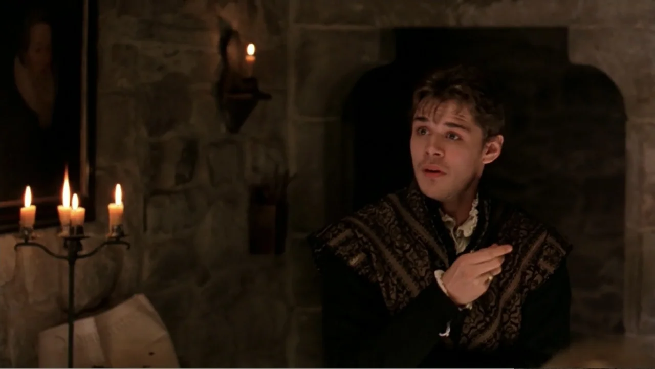 Elizabeth (1998) movie still: medium — A dimly lit room with heavy stone walls and a stone fireplace archway. A man in period clothing sits beside a…
