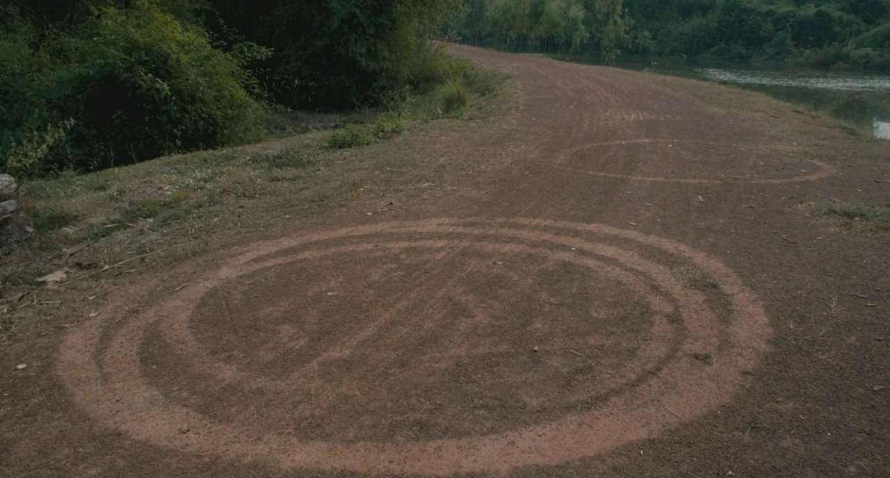 Uncle Boonme Who Can Recall His Past Lives (2010) movie still: wide — A dirt track runs alongside a river lined with dense green vegetation. Two…