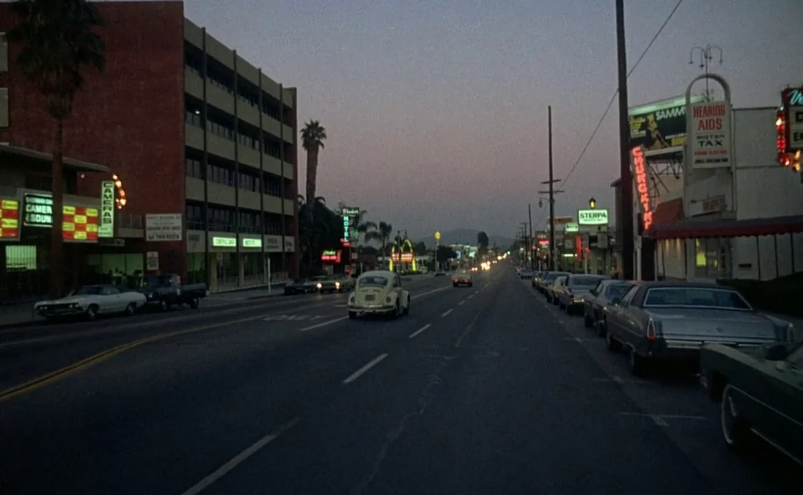 F for Fake (1973) movie still: establishing — A suburban street lined with low-rise commercial buildings and neon signage during dusk. A yellow…