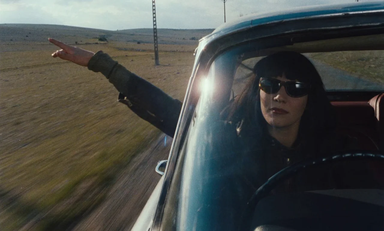 Until The End Of The World (1991) movie still: medium — A woman drives a vintage car along a dry, grassy, sparsely populated rural road. She has one…
