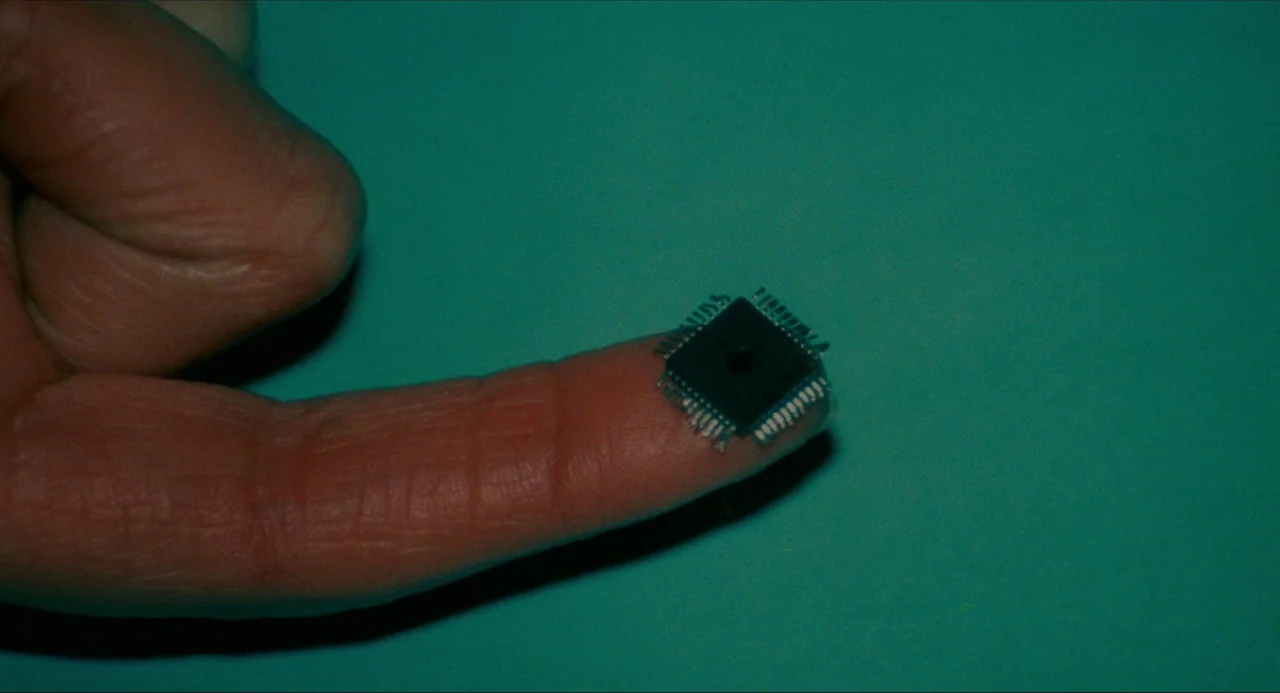 True Stories (1986) movie still: closeup — A close-up view shows a person's index finger against a solid teal-colored backdrop. A small square…
