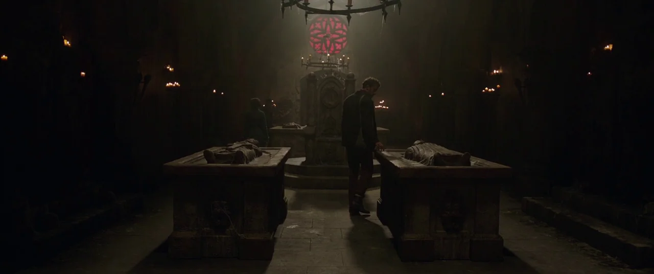 The Nun (2018) movie still: wide — A dark stone crypt features rows of elevated stone sarcophagi and arched walls. A man stands between the central…