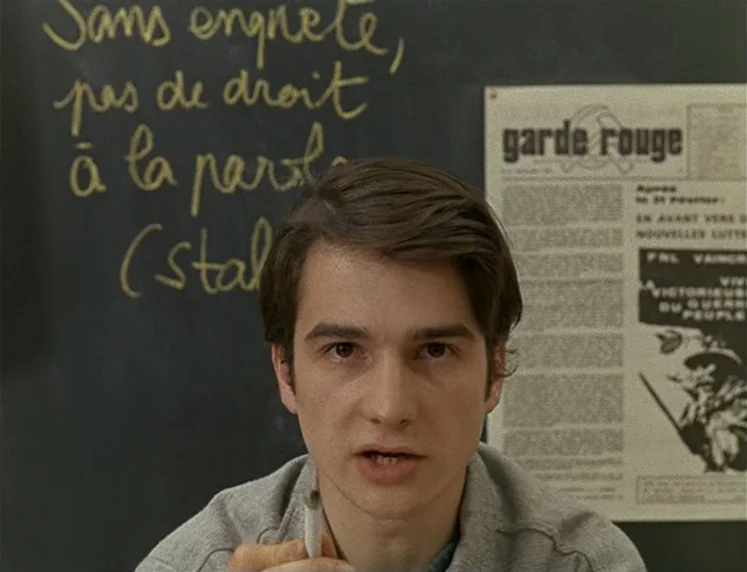 La Chinoise (1967) movie still: medium — A man stands in front of a blackboard covered in handwritten French text. A newspaper clipping titled Garde…