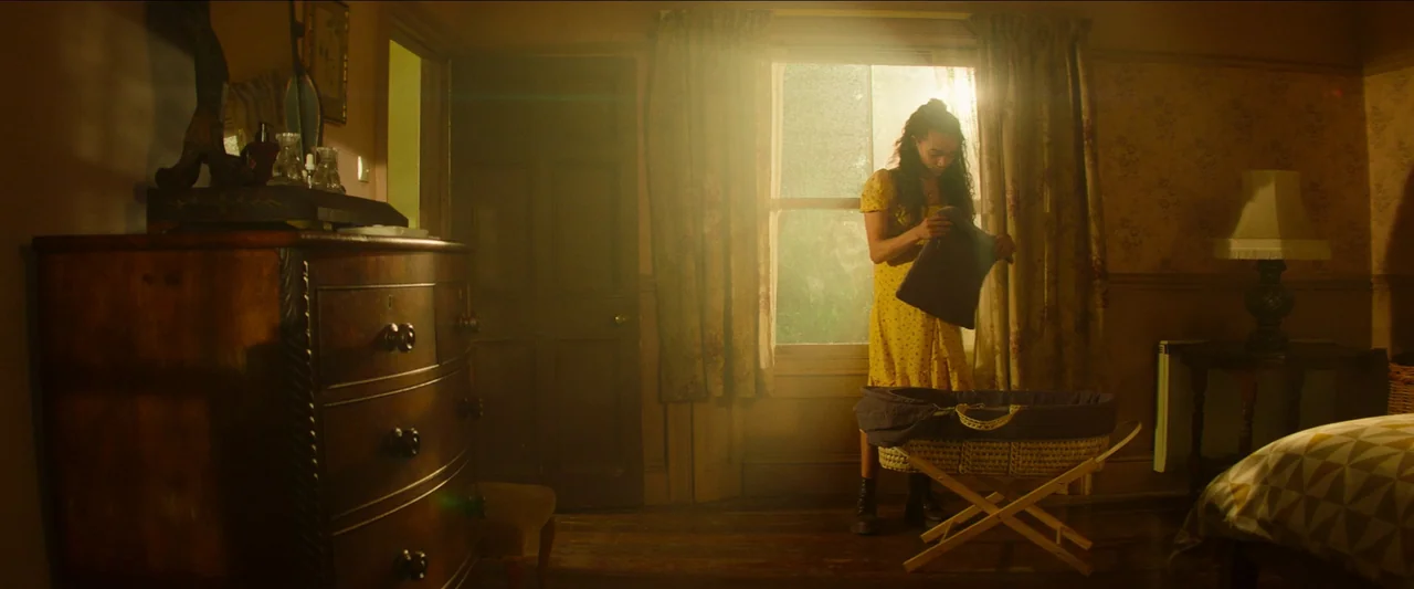 Unwelcome (2022) movie still: medium — A sunlit room features a dark wood dresser and a wicker bassinet near a window. A woman stands by the window,…