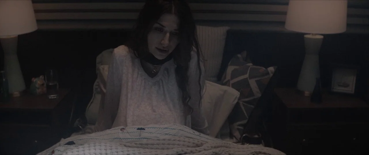 Anything For Jackson (2020) movie still: medium — A woman sits upright on a bed in a dimly lit, modern room. She looks downward while resting in the…