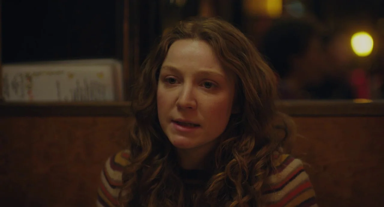 Private Life (2018) movie still: closeup — A dimly lit diner booth at night with a wood-paneled wall backdrop. A young woman with auburn hair sits…
