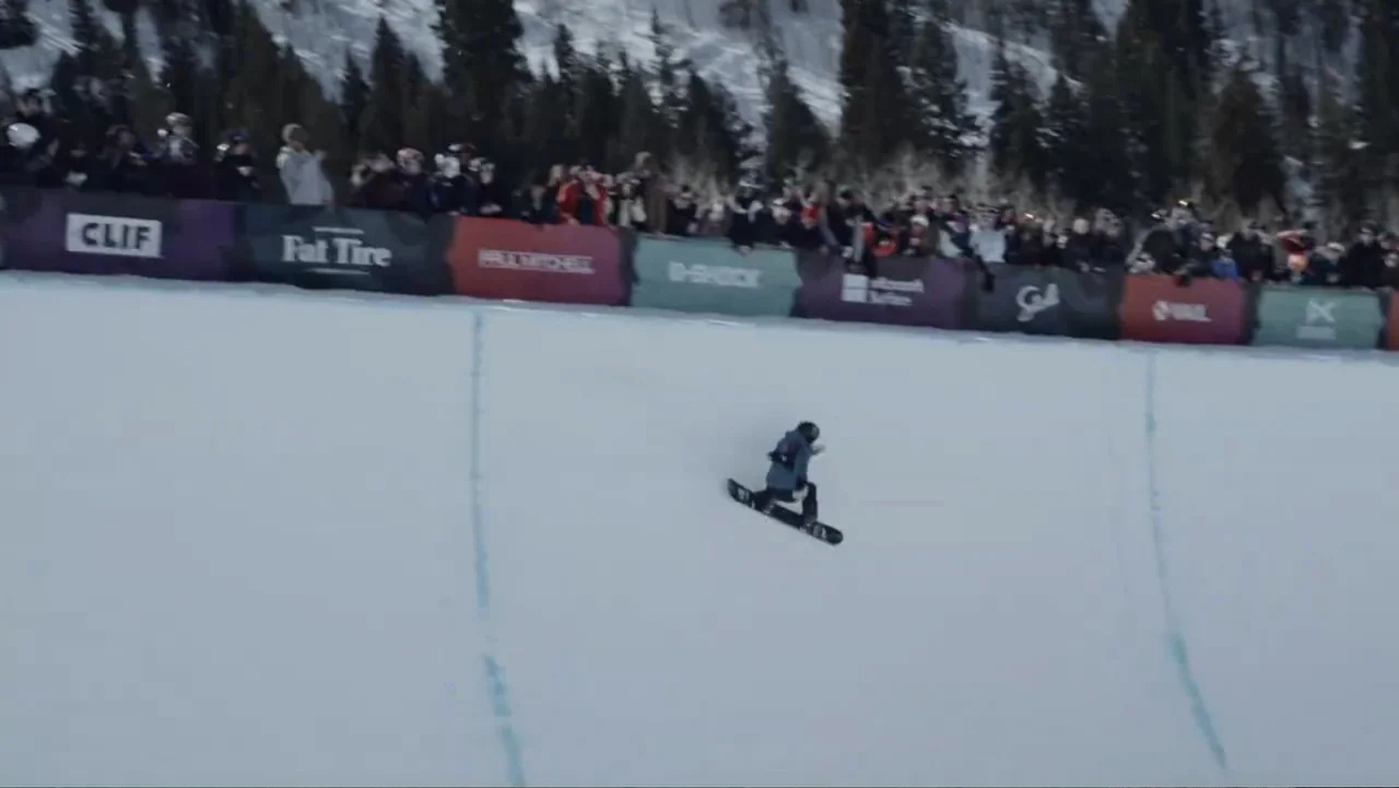 Dear Rider: The Jake Burton Story (2021) movie still: wide — A snowy halfpipe venue features a row of colorful sponsor banners against a treeline. A…