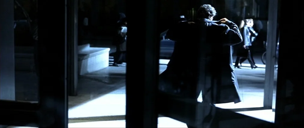 The Insider (1999) movie still: medium — A glass doorway separates a dark interior space from a brightly lit concrete sidewalk. A man in an overcoat…