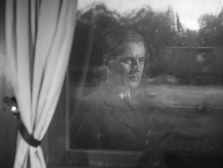 A Lesson In Love (1954) movie still: medium — A man looks through a window from inside a train car with curtains visible. The man is reflected in…