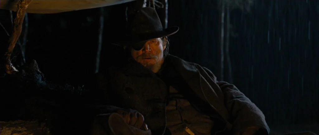 True Grit (2010) movie still: medium — An elderly man in a wide-brimmed hat sits outdoors during a dark rainstorm. He rests beneath a makeshift…