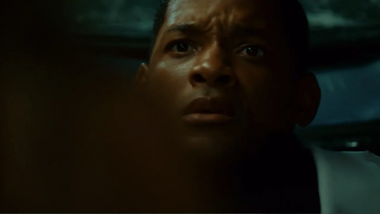 Hancock (2008) movie still: closeup — A dimly lit interior space with shadows obscuring most background details. A man stares intensely at an object…