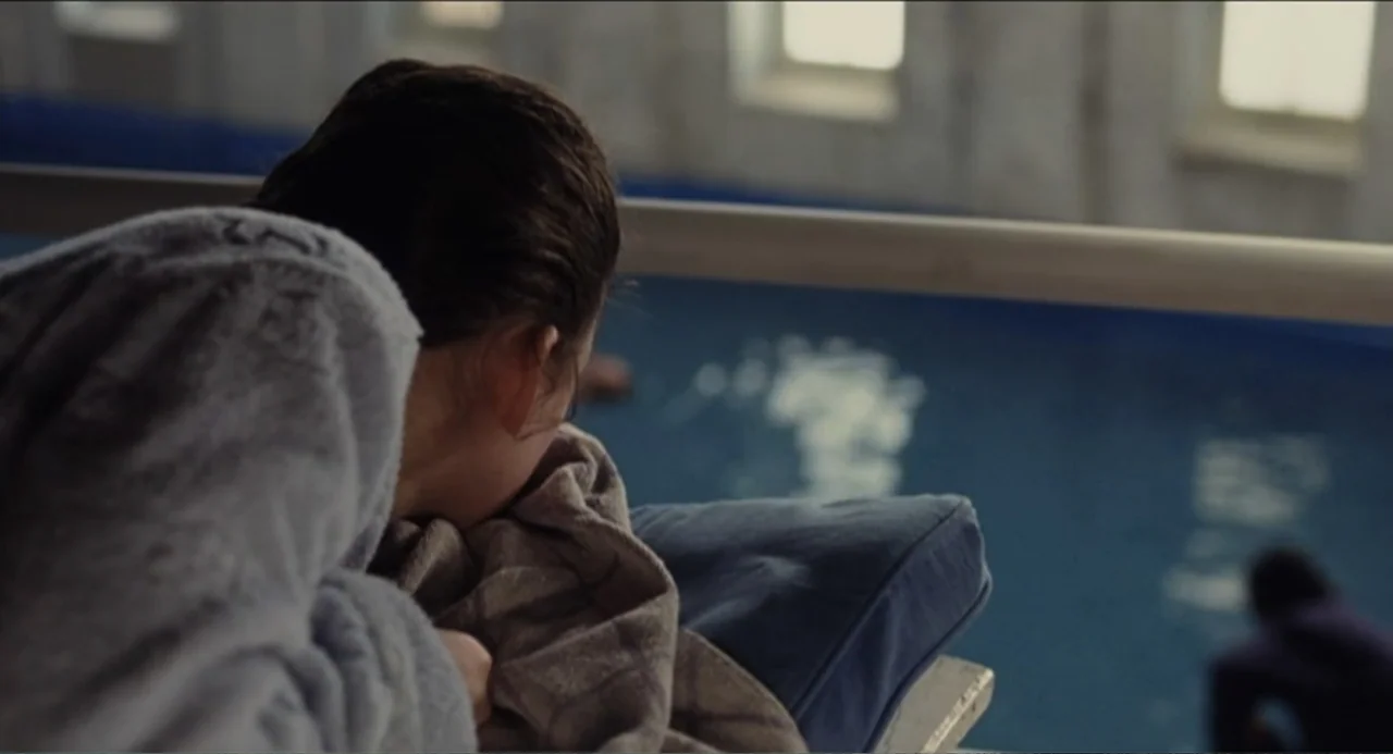 The Holy Girl (2004) movie still: medium — An indoor swimming pool area features blue water and white paneled walls with windows. A person wearing a…