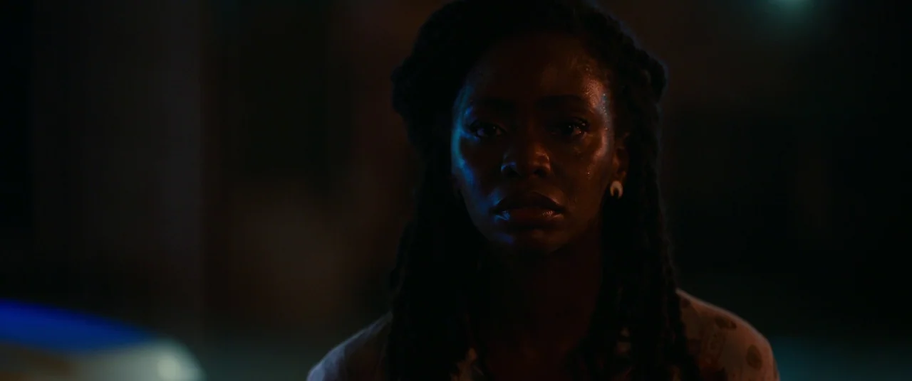 Candyman (2021) movie still: closeup — A dark outdoor setting at night illuminated by contrasting cool and warm light sources. A woman with long…