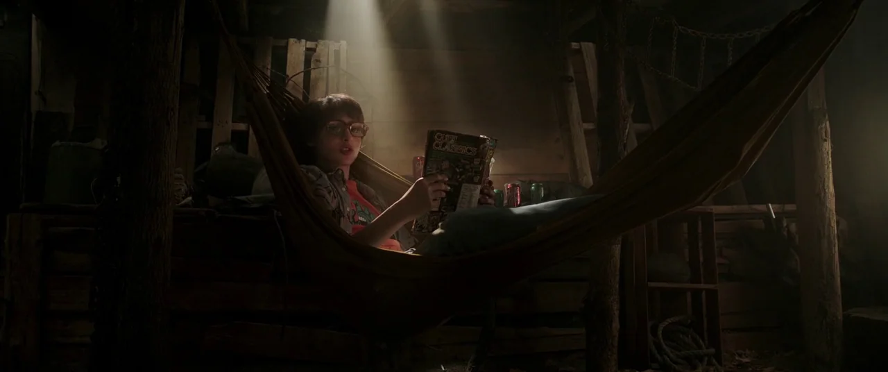 It Chapter Two (2019) movie still: medium — A dark wooden interior filled with shadows and rough timber support structures. A teenager rests in a…