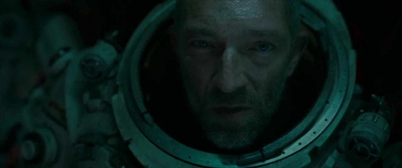 Underwater (2020) movie still: closeup — A close-up of a man inside a pressurized spacesuit with a metal collar. The lighting is dark and tinged…