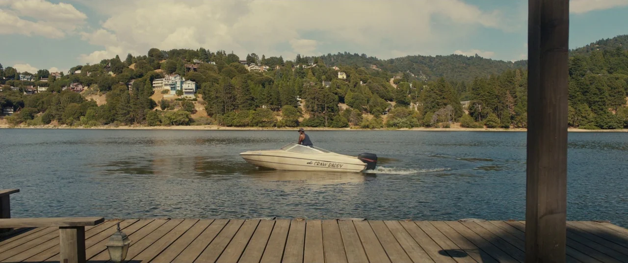 Us (2019) movie still: wide — A calm lake is framed by a wooden dock in the immediate foreground. A small motorboat named Craw Daddy travels across…