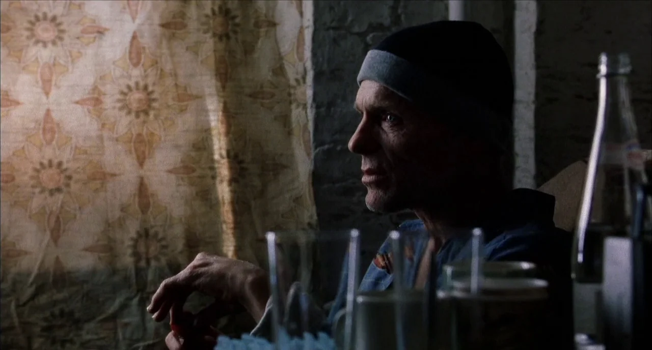 The Hours (2002) movie still: medium — A dimly lit room features a patterned curtain hanging against a rough textured wall. An older man sits in…