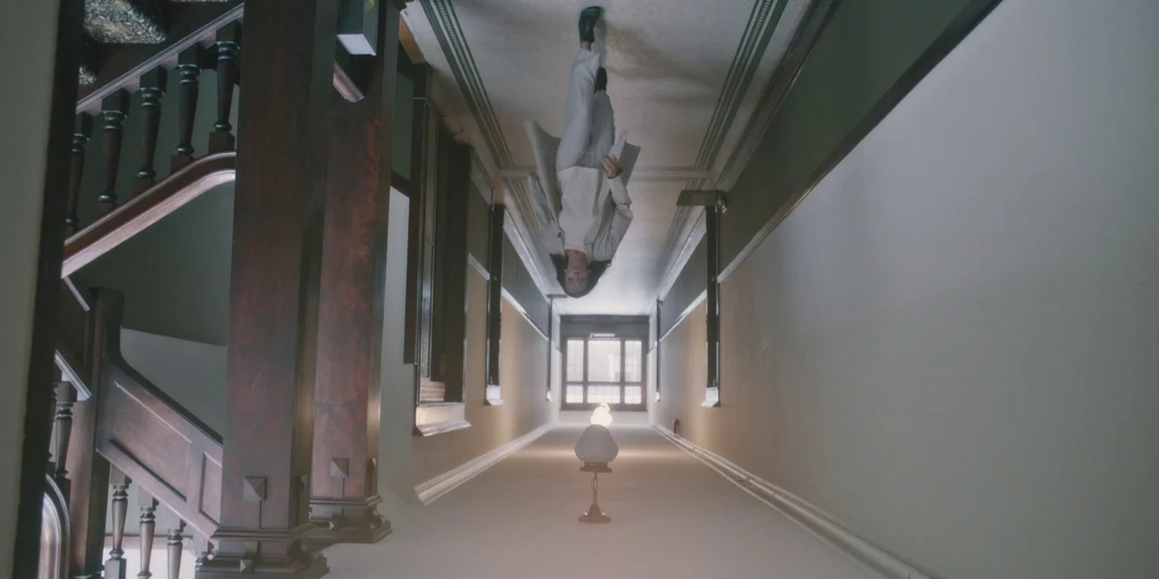 Hereditary (2018) movie still: wide — A long, minimalist hallway with wood paneled columns and white walls is shown upside down. A woman walks…