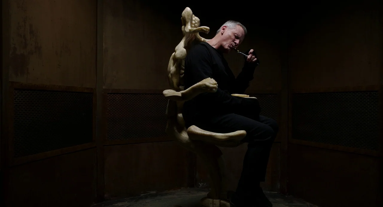 Crimes of the Future (2022) movie still: medium — A man sits on a bone-like sculpture chair in a dimly lit, walled room. The man eats from a small…