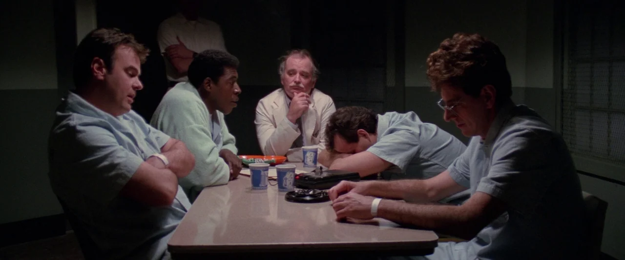 Ghostbusters 2 (1989) movie still: wide — A dimly lit room with industrial walls contains a central table and five men. The men sit around the table…