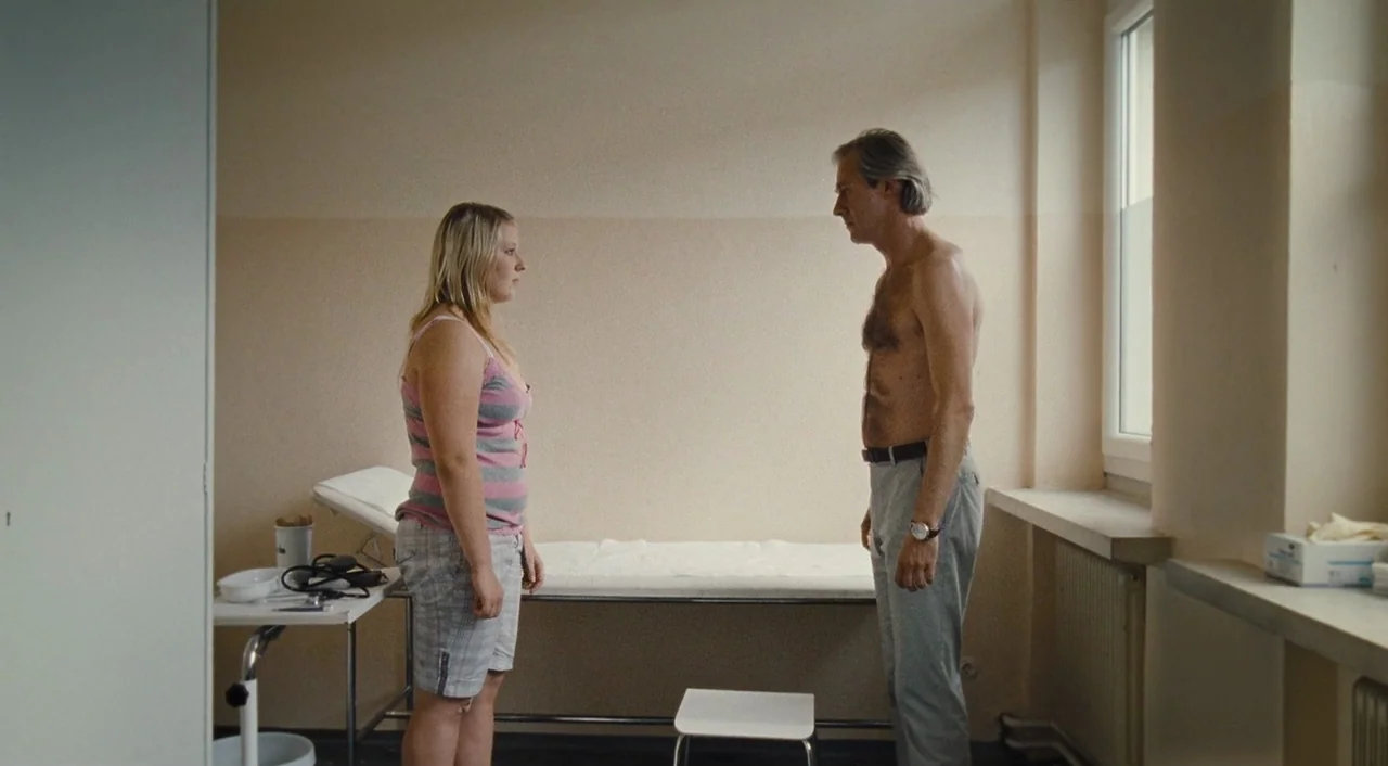 Paradise: Hope (2013) movie still: fullbody — A plain medical exam room contains a covered metal bed and a small rolling tray. A young woman and an…