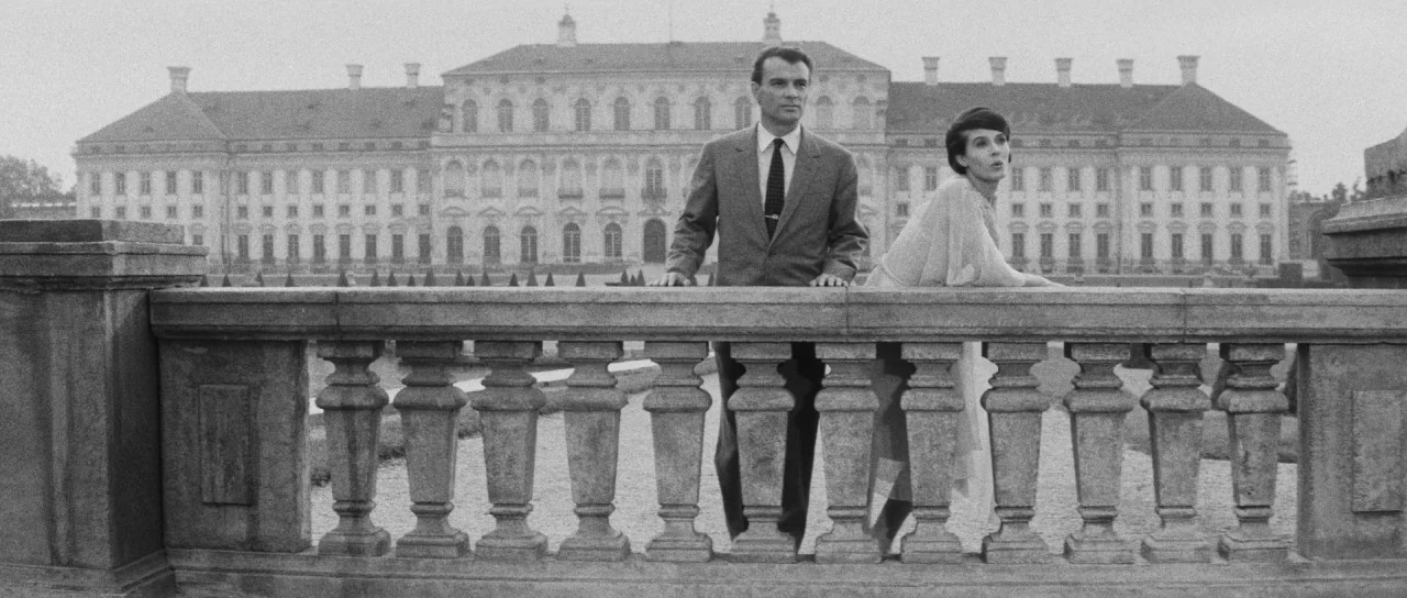 Last Year at Marienbad (1961) movie still: fullbody — A large baroque palace stands in the distance behind an ornate stone balustrade. A man and a…