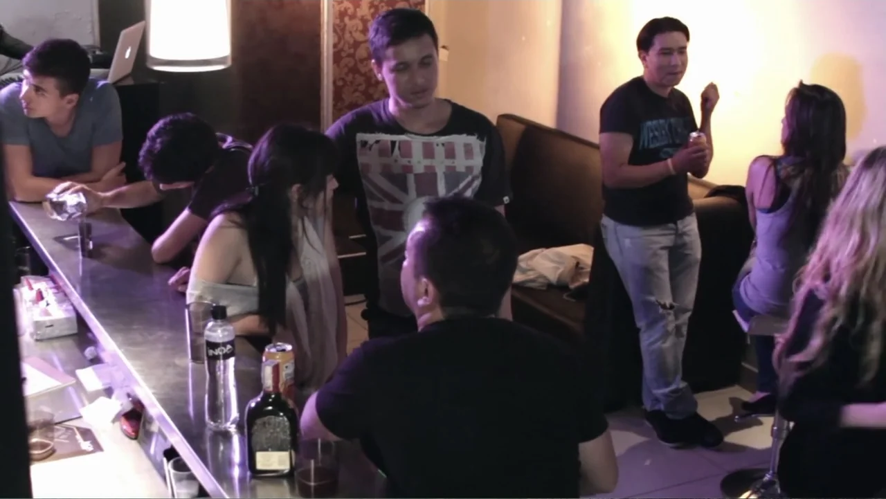 Aquel Septiembre (2018) movie still: wide — A modern bar counter area with metal surfaces and several patrons socializing together. A group of young…