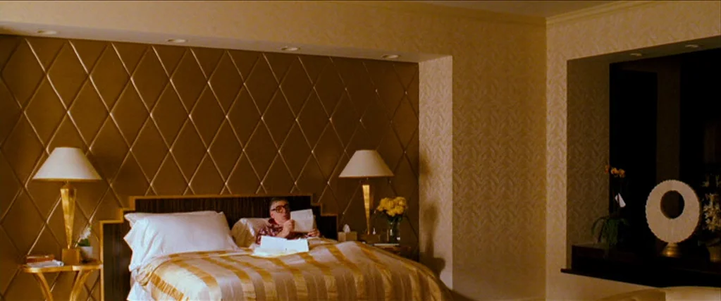 Ocean’s Thirteen (2007) movie still: wide — An opulent hotel suite features gold padded diamond-patterned walls and dual bedside lamps. An elderly…