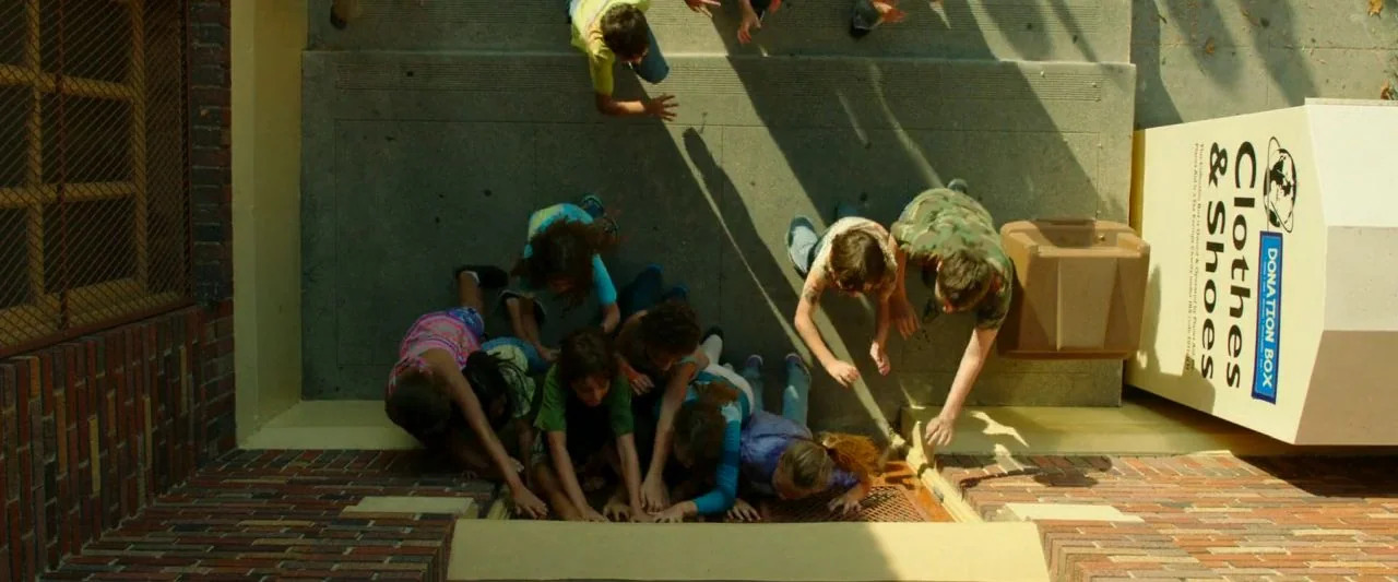 Cooties (2014) movie still: wide — A concrete sidewalk outside a brick building features a donation box. Multiple children huddle closely together…