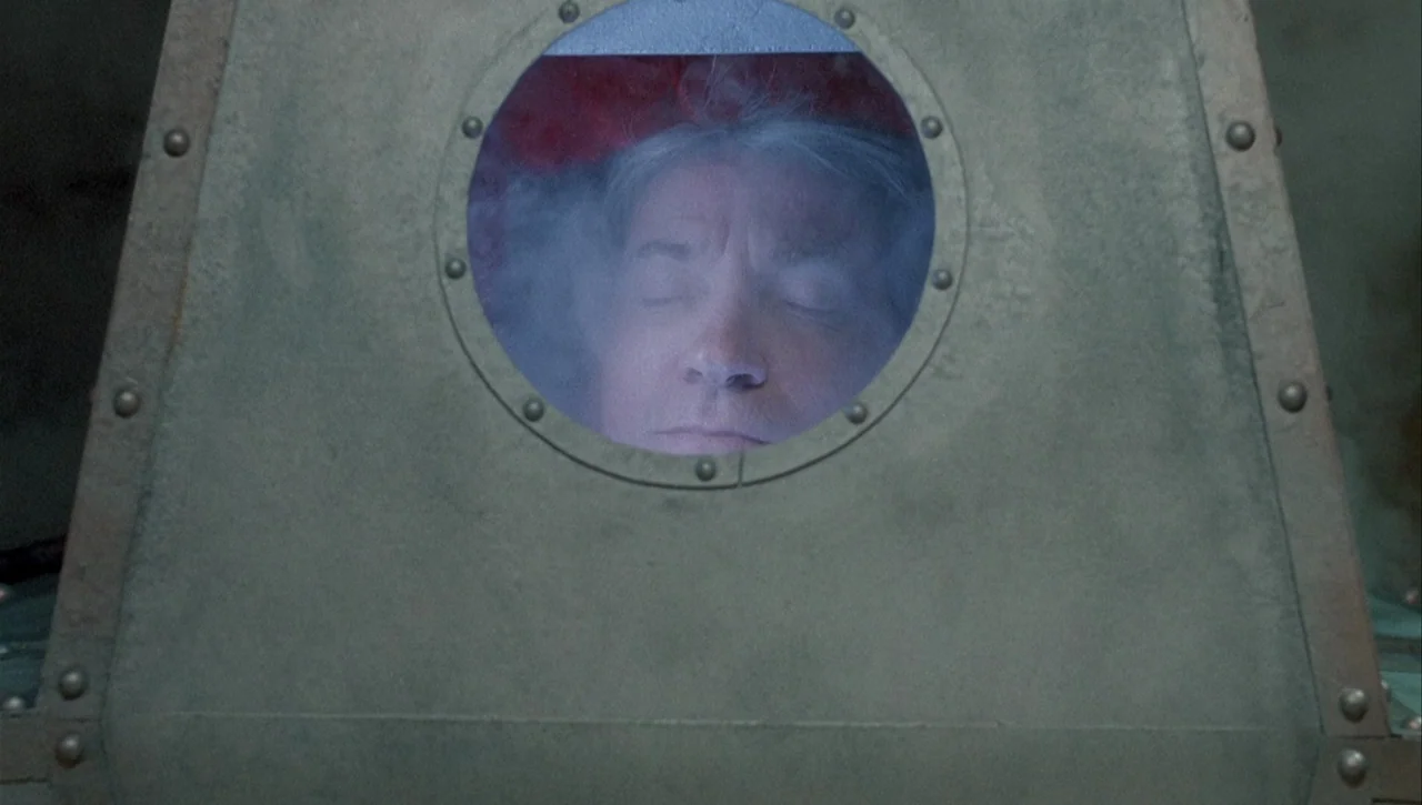 Necronomicon: Book of Dead (1993) movie still: closeup — A man's face is visible through the circular porthole of a metallic chamber. He lies with…