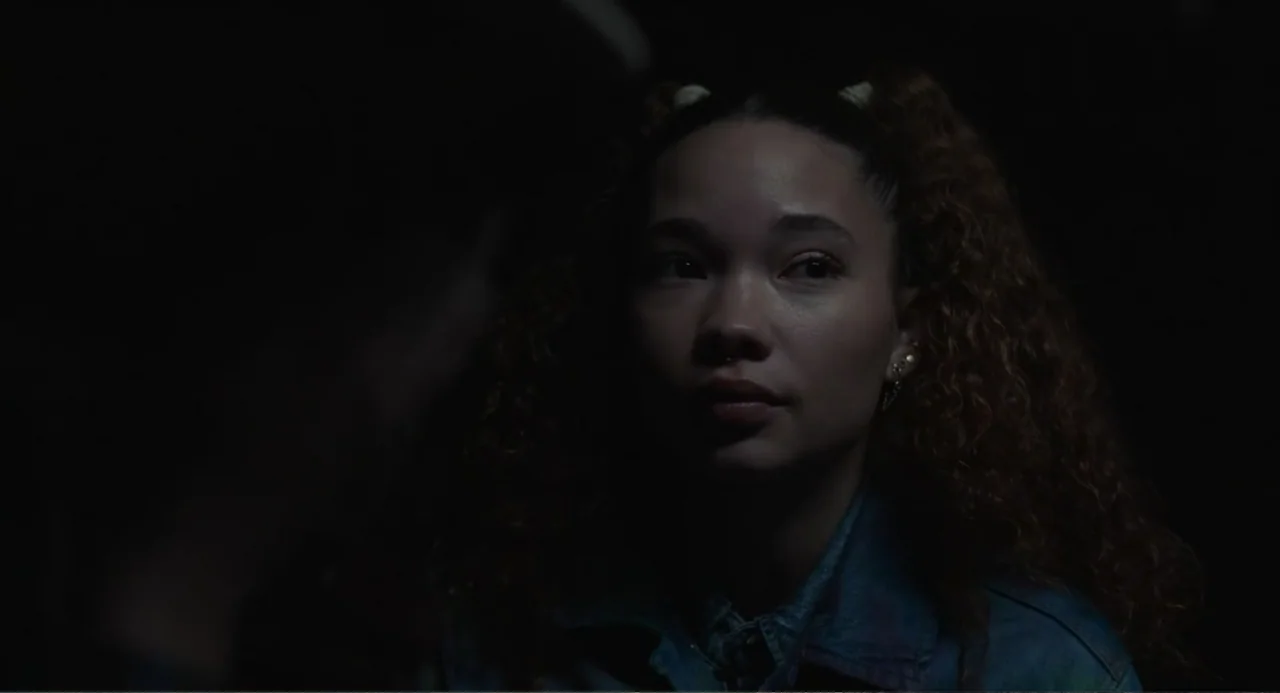 Salvation (2025) movie still: closeup — A dimly lit interior space characterized by heavy shadow and minimal background detail. A young woman with…