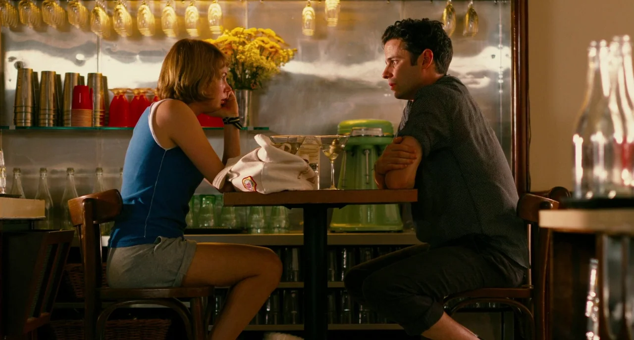Take This Waltz (2011) movie still: medium — A man and woman sit at a small metal table inside a diner. Yellow flowers sit in a glass vase between…