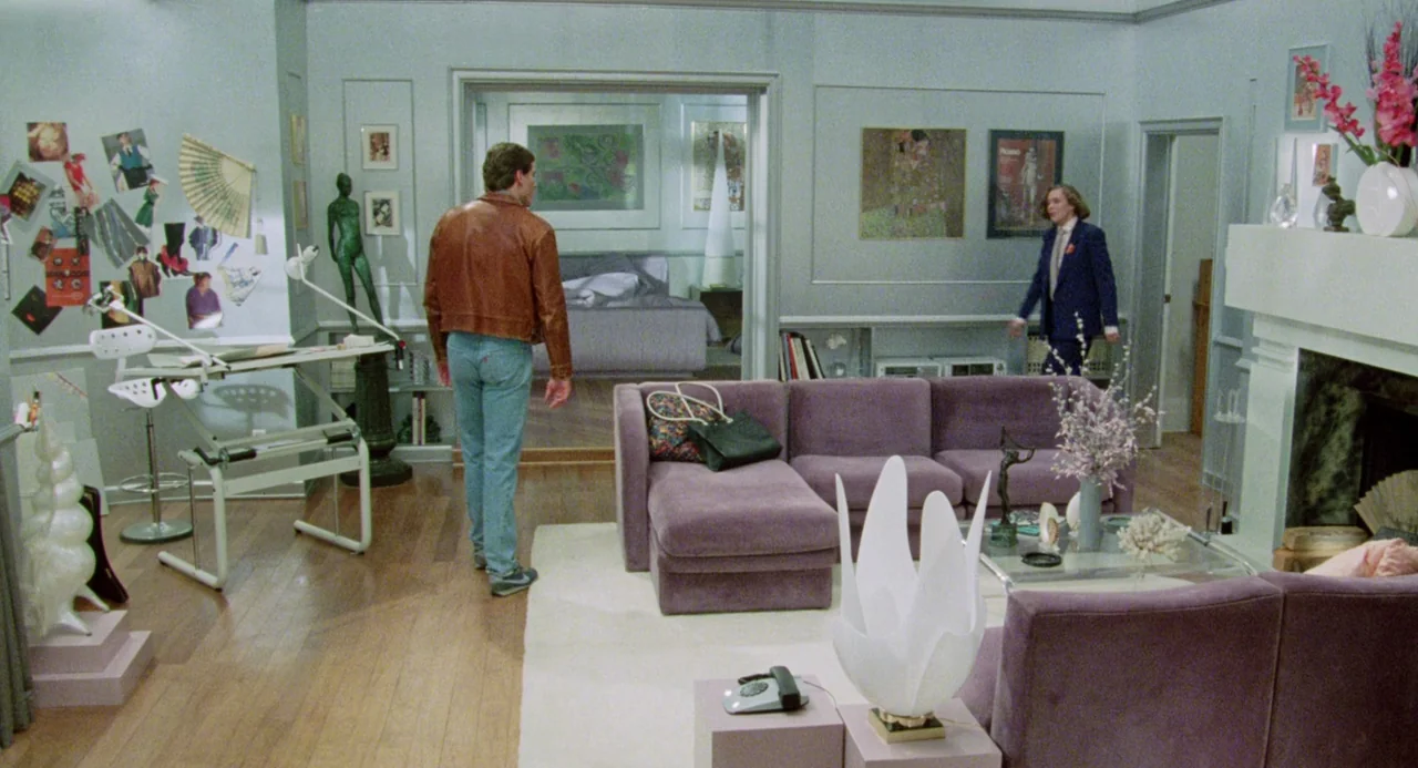 Crimes of Passion (1984) movie still: wide — A spacious 1980s living room features a purple sectional sofa and hardwood flooring. One man stands by…