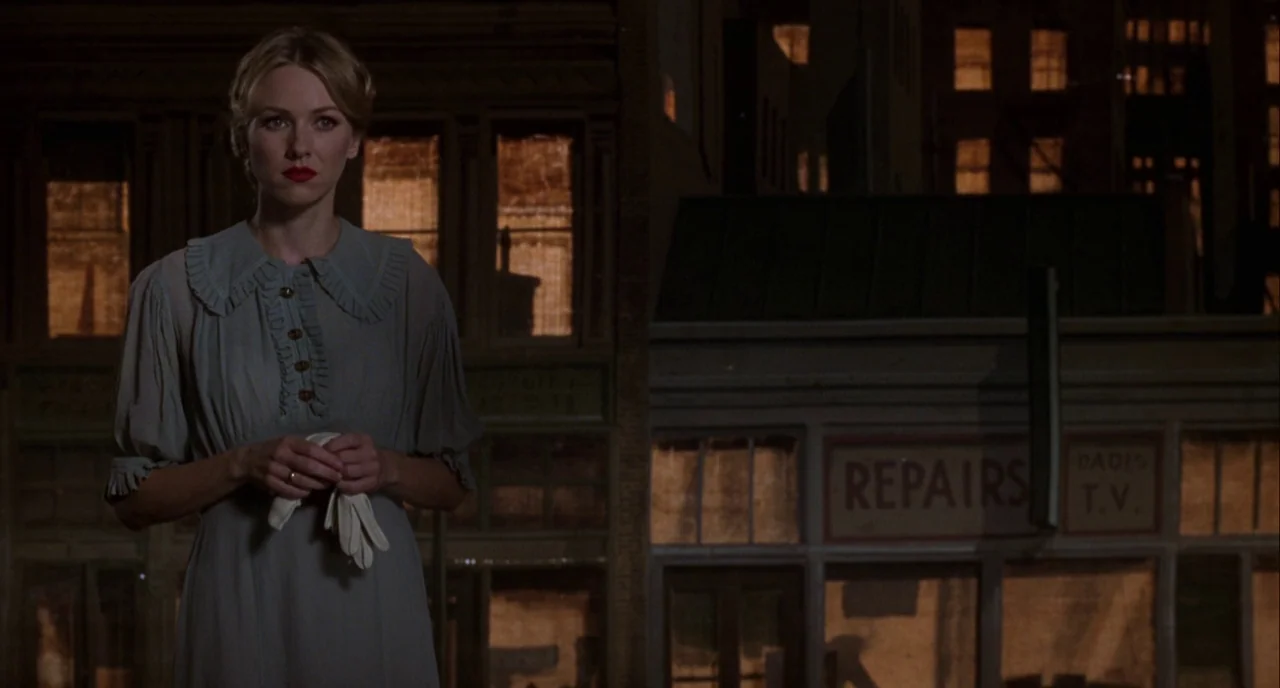 Mulholland Drive (2001) movie still: medium — A nighttime city street features a storefront with a repair shop sign in window. A woman stands in the…
