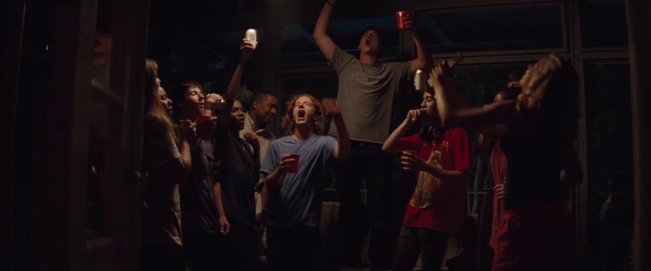 The Spectacular Now (2013) movie still: medium — A dimly lit glass-enclosed room at night holds a group of rowdy young adults. The group stands…